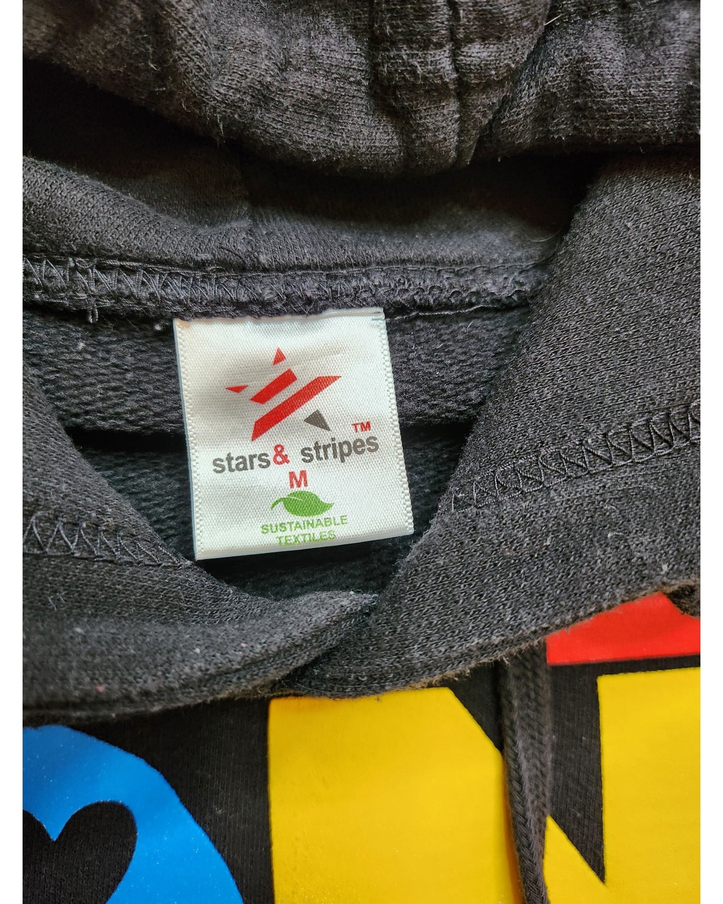 Close-up of a touched out mama clothing tag on the Upcycled Breastfeeding & Pumping Harry Styles Hoodie M. Vibrant red, blue, and yellow showcase its eco-friendly fashion and bold red star logo celebrating sustainable design.