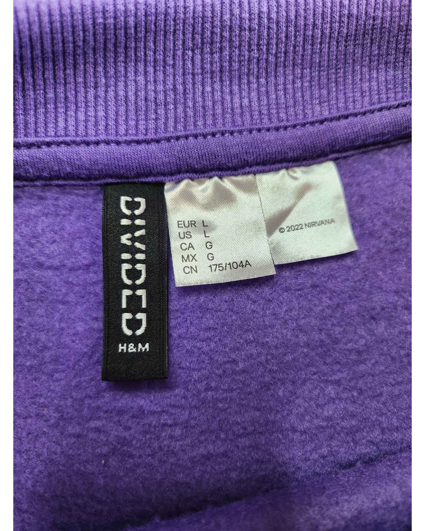 Close-up of clothing labels on a preloved purple fabric feature regional sizing and touched out mama. Another tag reads ©2022 NVIRANA. This upcycled piece, the Upcycled Breastfeeding & Pumping Nirvana Sweatshirt L, showcases a ribbed edge at the top.