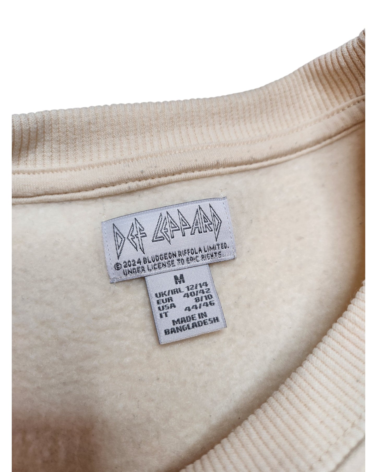 Close-up of a label inside the collar of an upcycled Def Leppard cream sweatshirt, part of the touched out mama line. The label displays size info (S/KR/USA/EU) and Made in Bangladesh.