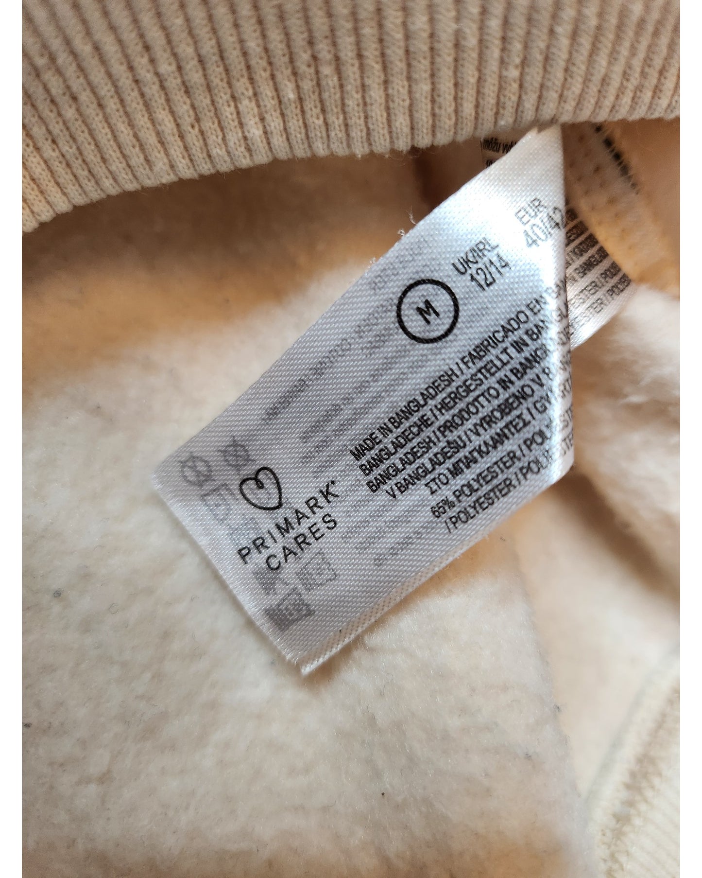 Close-up of a PRIMARK CARES label in a beige garment. The eco-friendly tag has multilingual care tips, indicating an M-sized polyester product named Upcycled Breastfeeding & Pumping Def Leppard Cream Sweatshirt by touched out mama, made in Bangladesh.