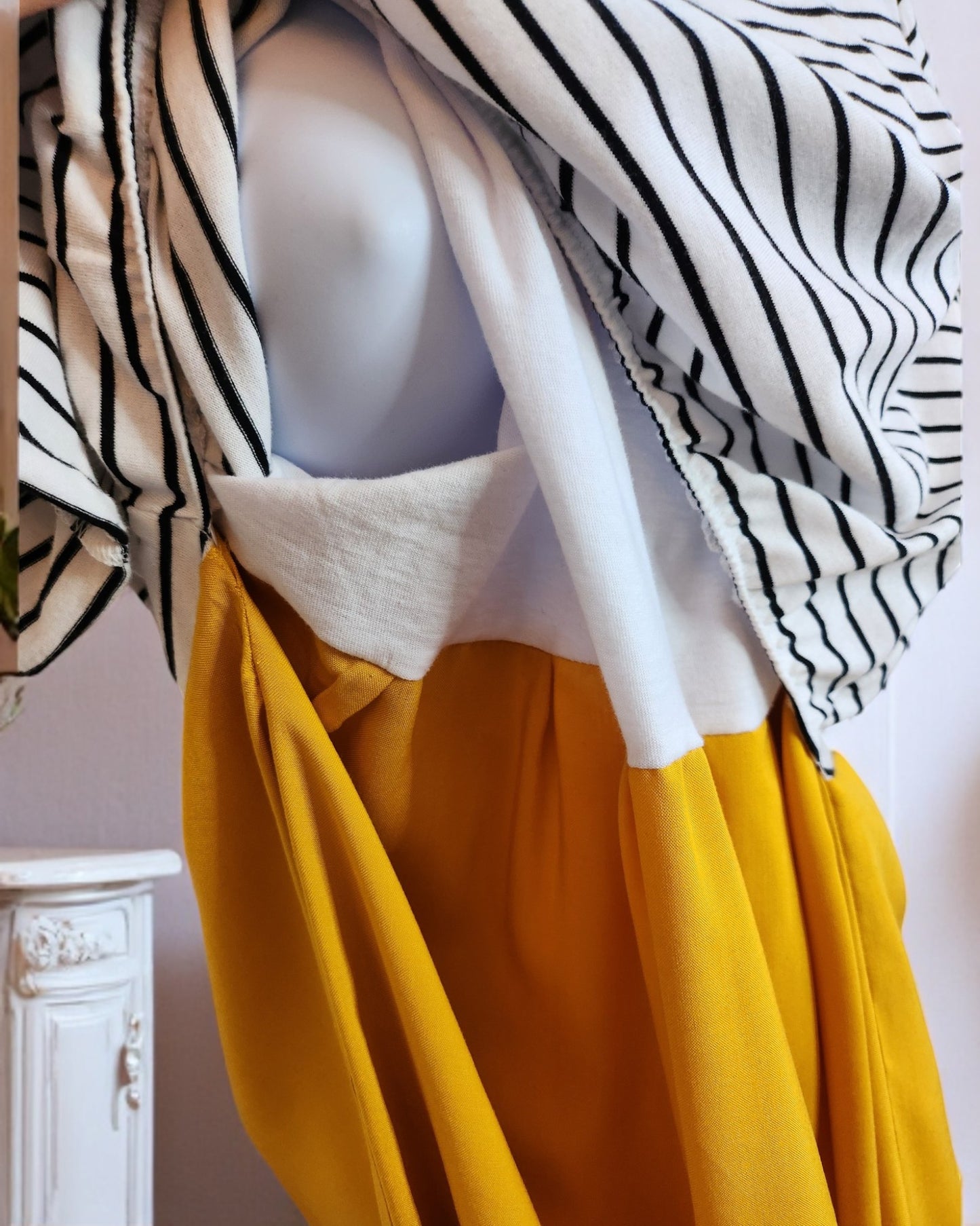 Close-up of a mannequin wearing the eco-friendly Upcycled Breastfeeding & Pumping Stripe Ochre Maxi Dress by touched out mama, combining a striped white and black top with a yellow bottom. A decorative white cabinet is partially visible beside the mannequin.
