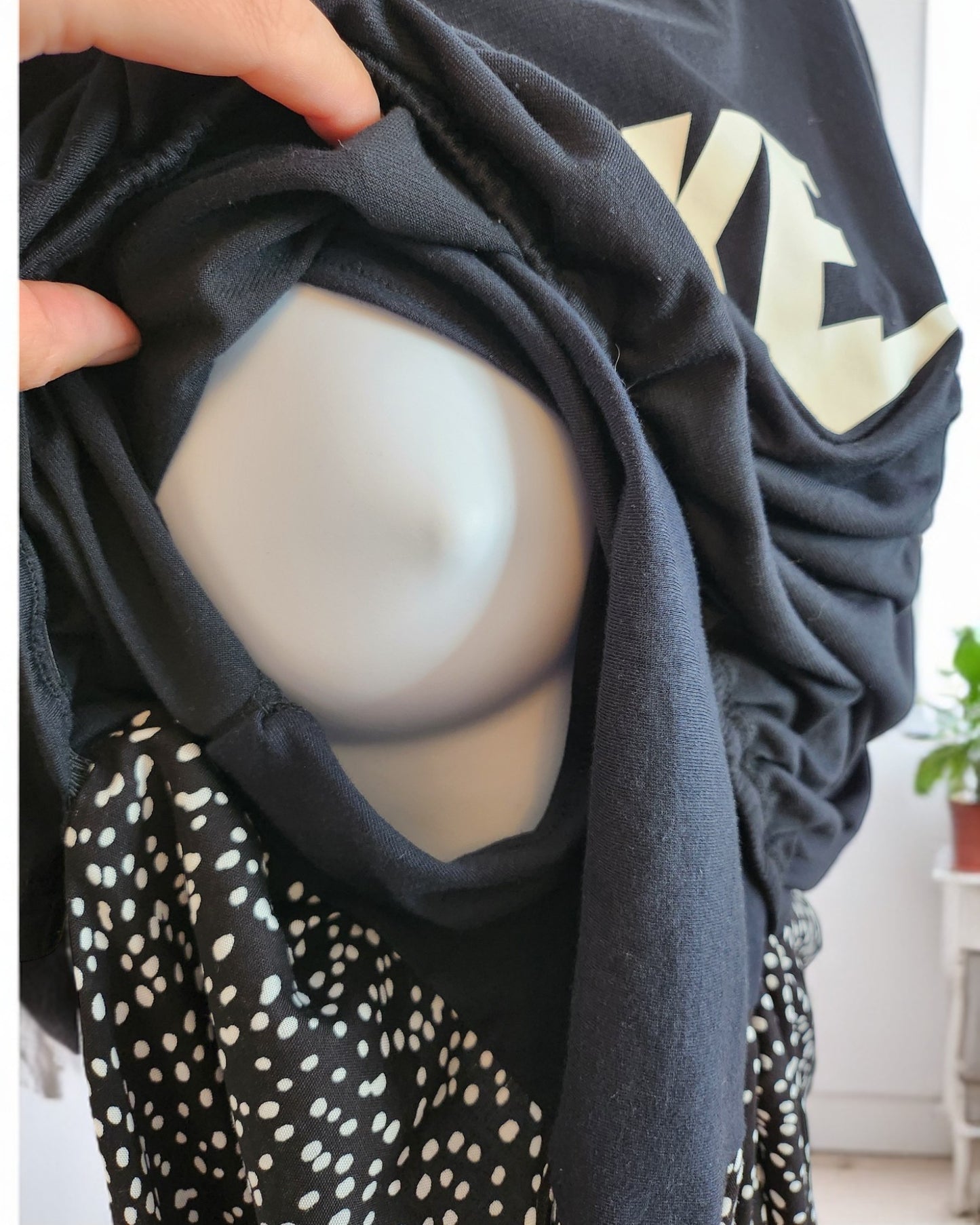 A mannequin head wrapped in an upcycled black NIKE sweatshirt from the touched out mama collection reveals white lettering and polka dot fabric. A hand adjusts this preloved maternity item, and a potted plant is seen in the background.