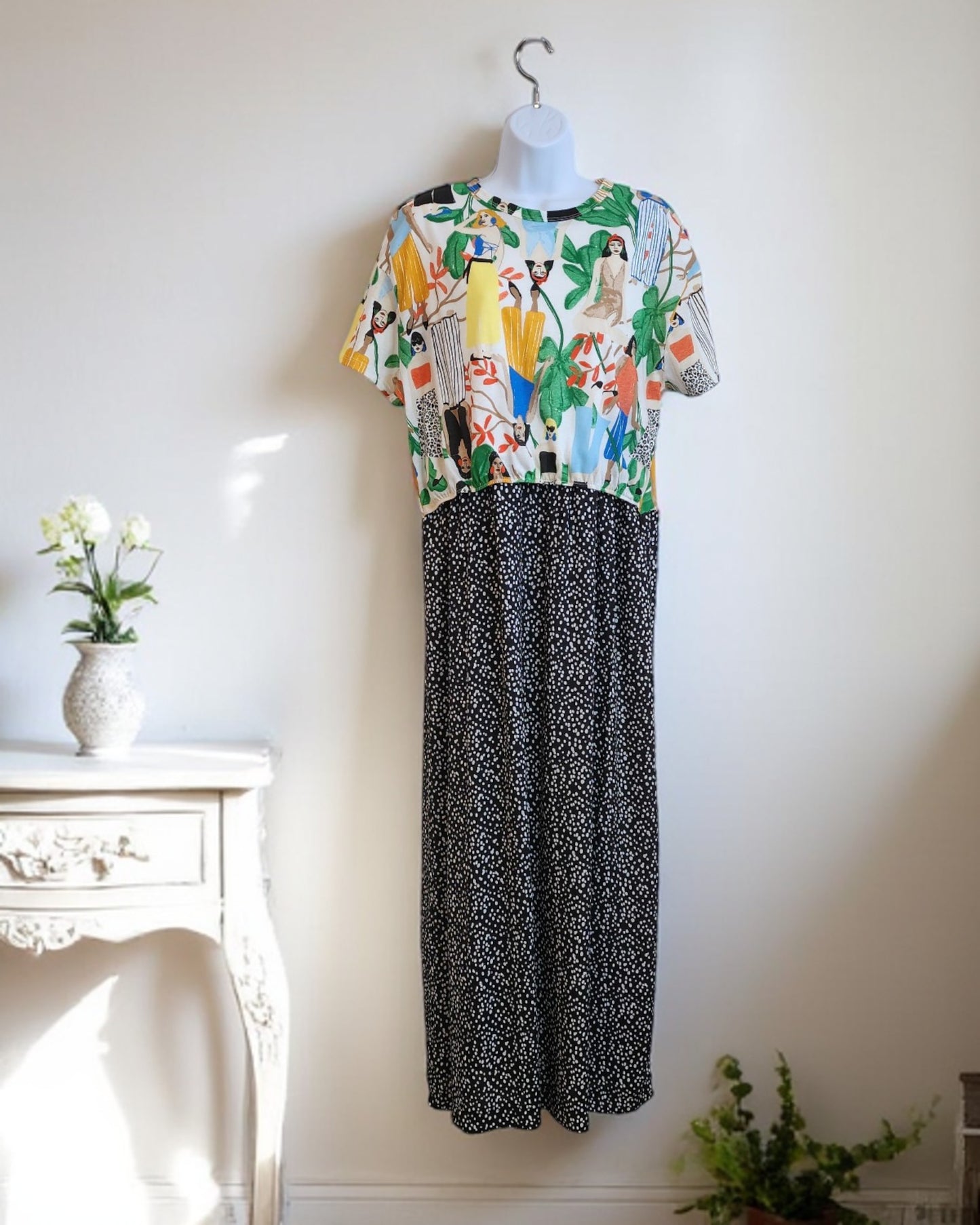 The touched out mama Upcycled Breastfeeding & Pumping ZARA Tropical Maxi Dress M/L features a vibrant abstract-patterned top and a long black skirt with white dots. It pairs beautifully with a decorated table holding a white vase and green plant against a neutral wall.