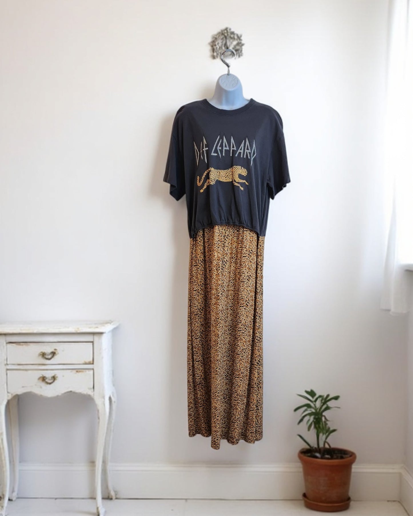 A mannequin showcases the Upcycled Breastfeeding & Pumping Def Leppard Maxi Dress M/L by touched out mama, embodying preloved style. Positioned against a plain white wall, it is paired with a small vintage-style table and a potted plant.