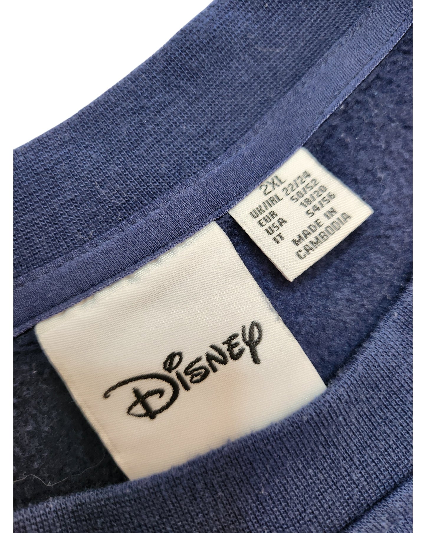 Close-up of the neckline on a blue Primark Upcycled Breastfeeding & Pumping Eeyore Disney Sweatshirt (2XL), showing two tags: one with the Disney logo and one with size/care info for USA, UK, and EU—made in Cambodia with an eco-friendly focus.