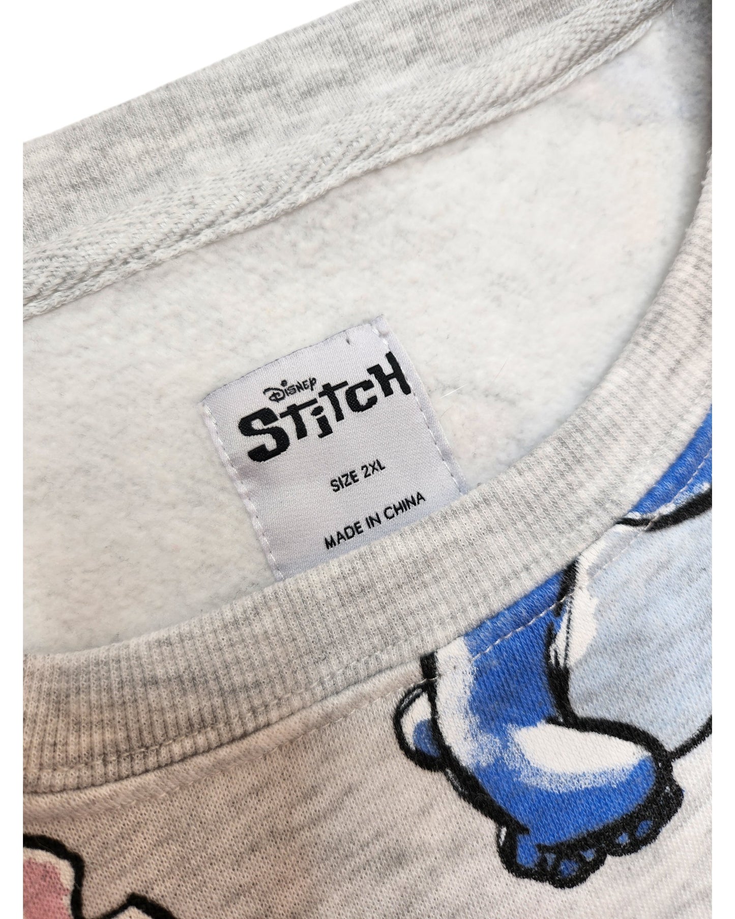 Close-up of a Primark Upcycled Breastfeeding & Pumping Stitch Disney Sweatshirt, 2XL. The gray shirt’s collar tag is shown, with soft fabric featuring part of the blue cartoon character Stitch. Preloved and made for comfort.