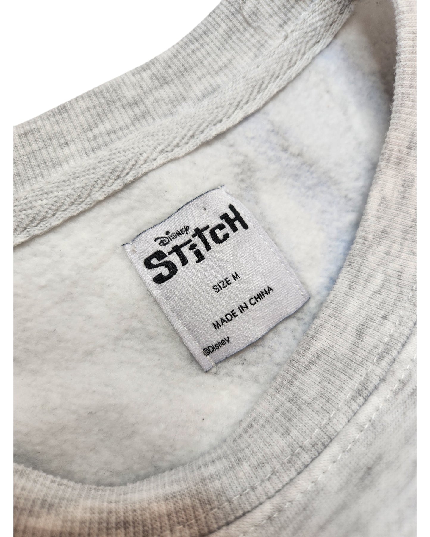Close-up of a white tag inside a light gray, upcycled touched out mama breastfeeding & pumping Disney Stitch sweatshirt (size M), made in China, sewn onto soft fleece-like fabric.