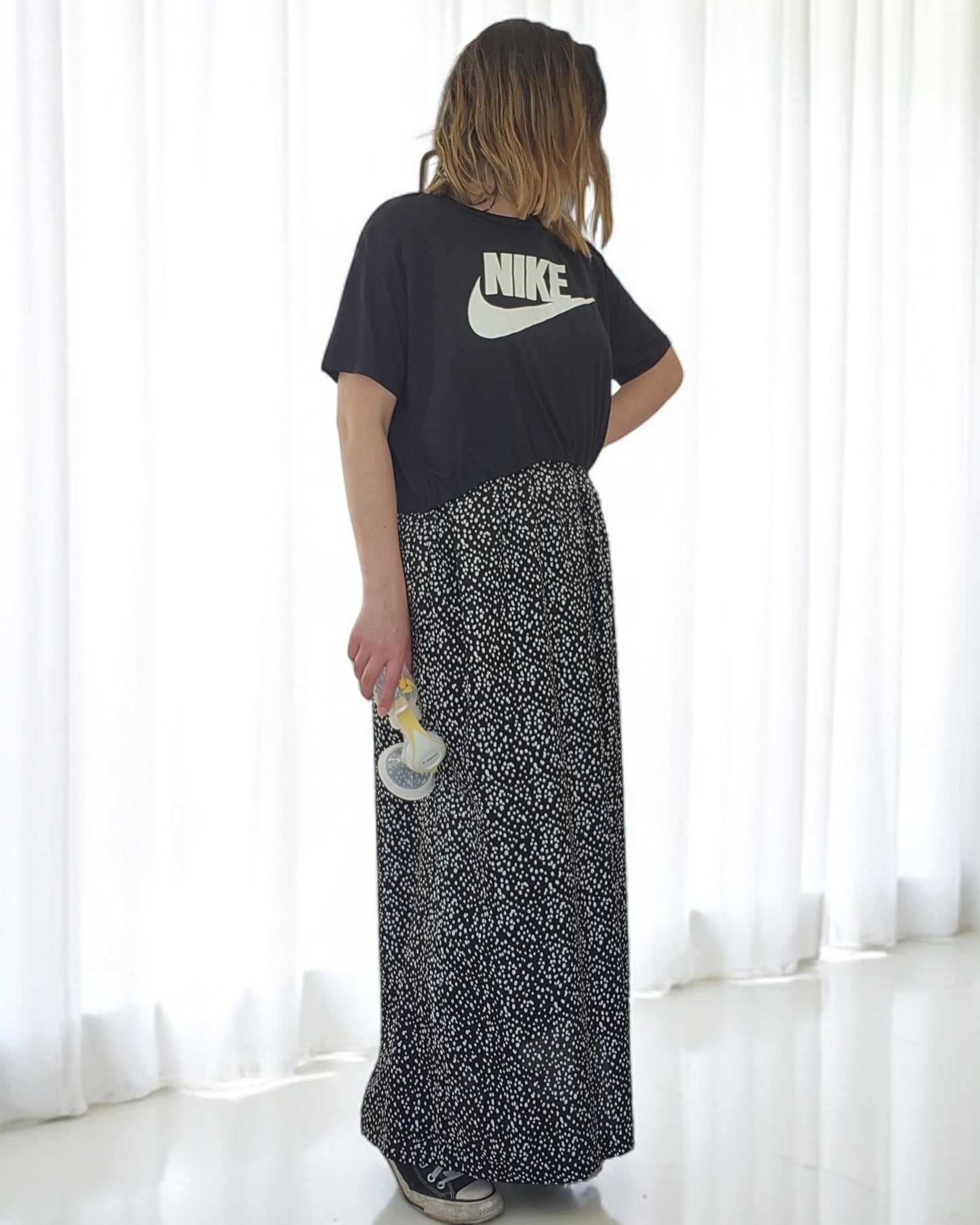 In front of white curtains, a person with shoulder-length hair wears the Upcycled Breastfeeding & Pumping NIKE Maxi Dress by touched out mama. The black and white patterned maxi dress and the breast pump emphasize the realities of nursing as their face turns away.