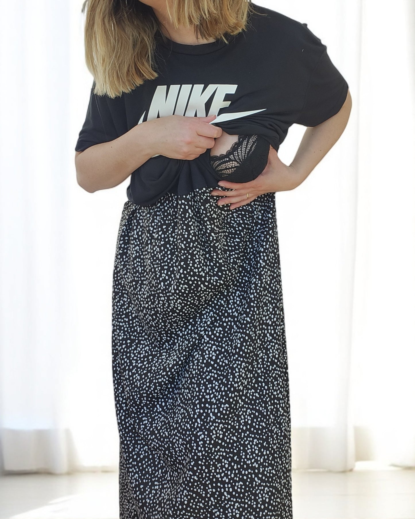 A person in a touched out mama Upcycled Breastfeeding & Pumping NIKE Maxi Dress M/L stands indoors, adjusting the black shirt at the waist. The background has white curtains and bright lighting, with the skirt featuring a long black and white dotted pattern.