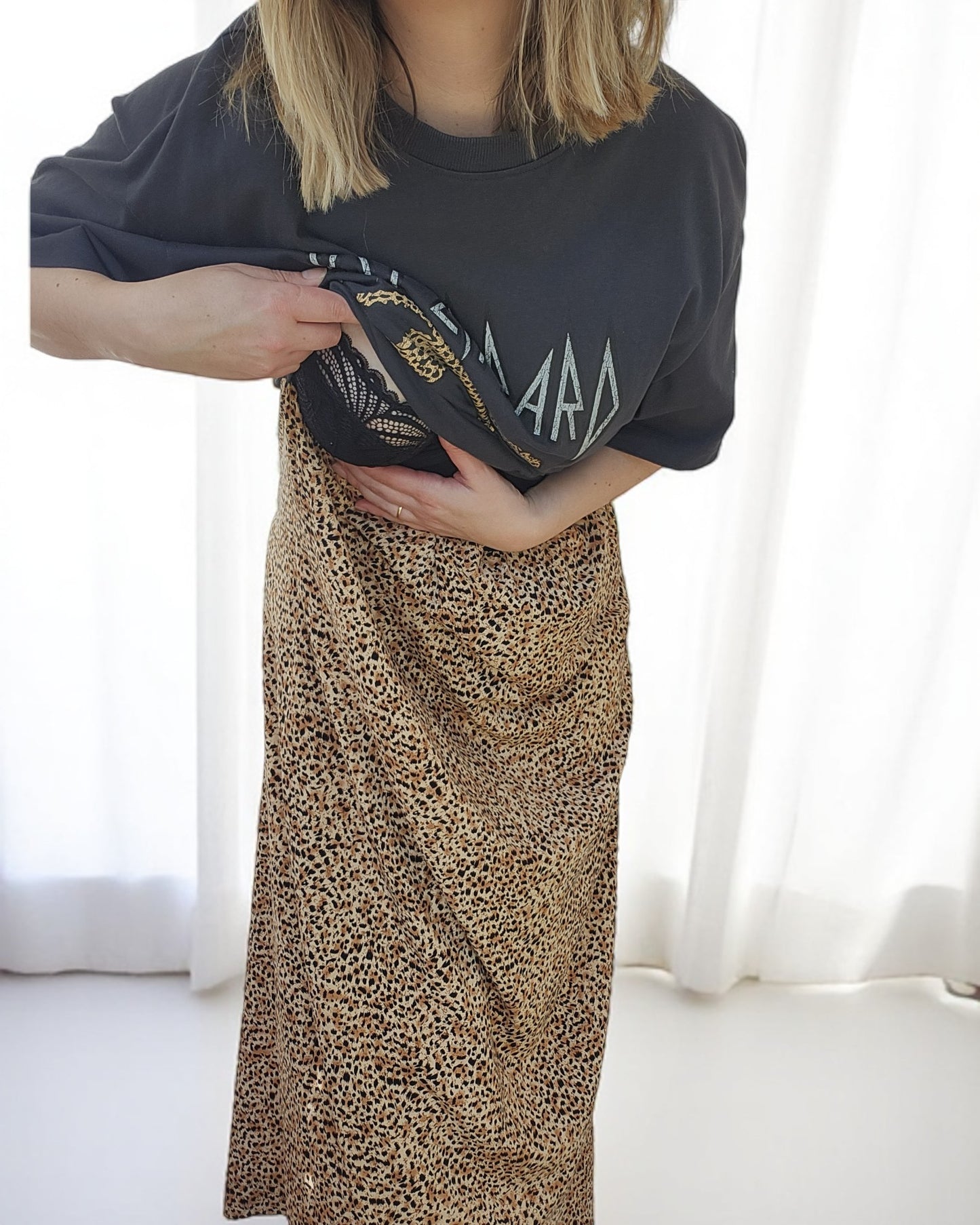 A light-haired person styled in an Upcycled Breastfeeding & Pumping Def Leppard Maxi Dress by touched out mama subtly reveals a leopard print bra, embodying upcycled fashion against white curtains.