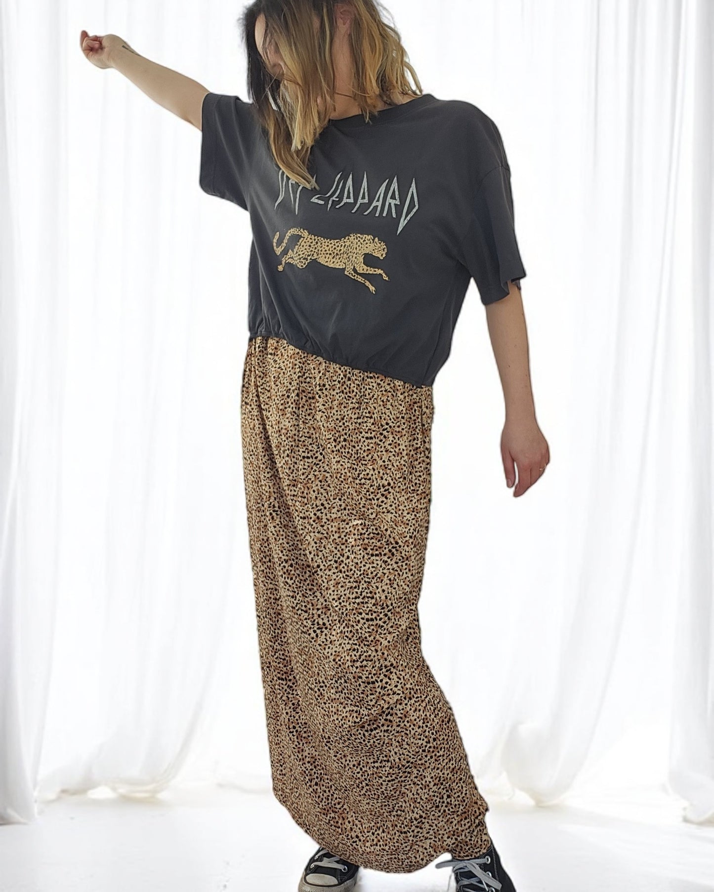 Against a white backdrop, someone showcases the Upcycled Breastfeeding & Pumping Def Leppard Maxi Dress from touched out mama. With light hair and an arm raised slightly overhead, they effortlessly embody the essence of sustainable fashion.