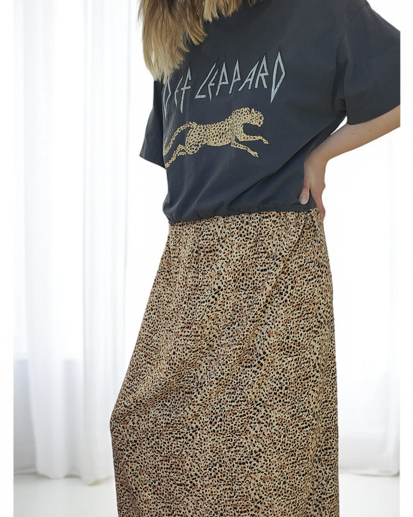 A person wearing a preloved black Def Leppard nursing & pumping maxi dress in a breathtaking leopard print by touched out mama stands confidently against a light-colored curtain backdrop. The upcycled fashion ensemble exudes uniqueness and style.