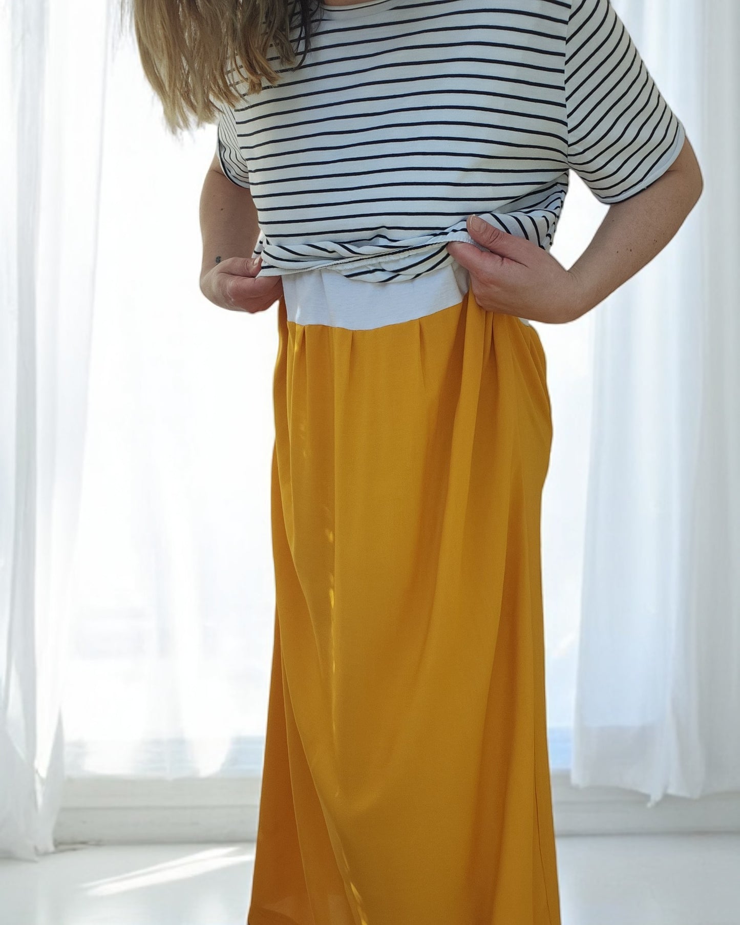 In front of a sunlit white curtain, someone wearing the touched out mama Upcycled Breastfeeding & Pumping Stripe Ochre Maxi Dress M/L is adjusting their yellow skirt.