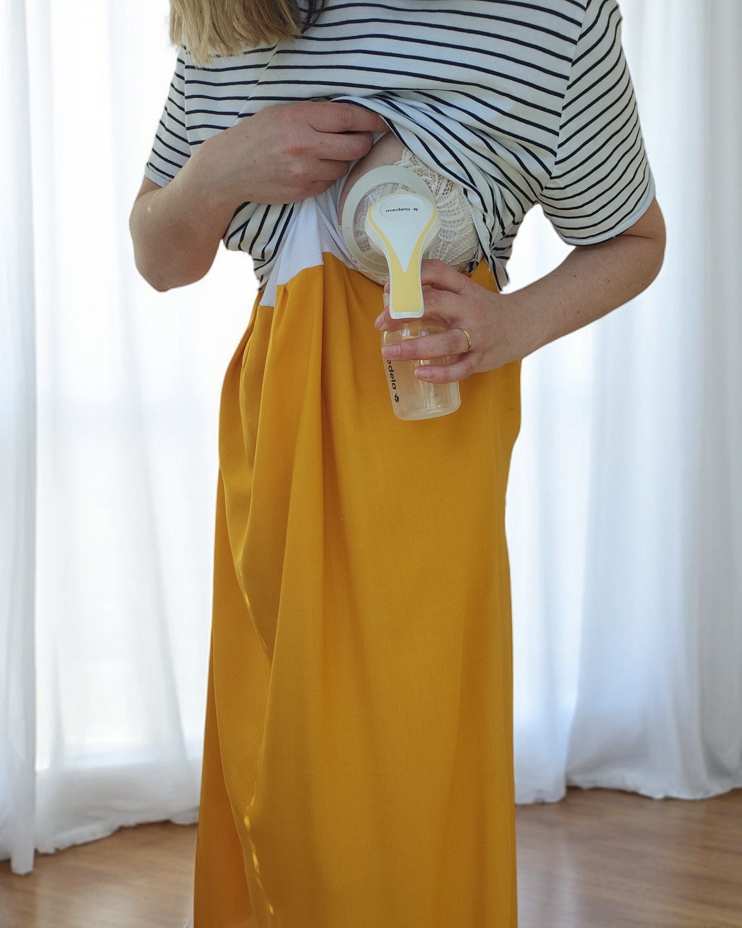 A person in eco-friendly fashion wears a touched out mama Upcycled Breastfeeding & Pumping Stripe Ochre Maxi Dress (M/L) and discreetly uses a manual breast pump under their dress, standing before white curtains.