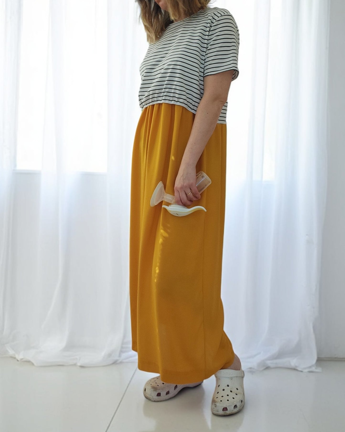 In a minimalist room with sheer white curtains, a person dons an Upcycled Breastfeeding (Nursing) & Pumping Stripe Ochre Maxi Dress by touched out mama, embodying eco-friendly fashion. They hold a pair of shoes and wear white slip-on clogs, enhancing the sustainable style.