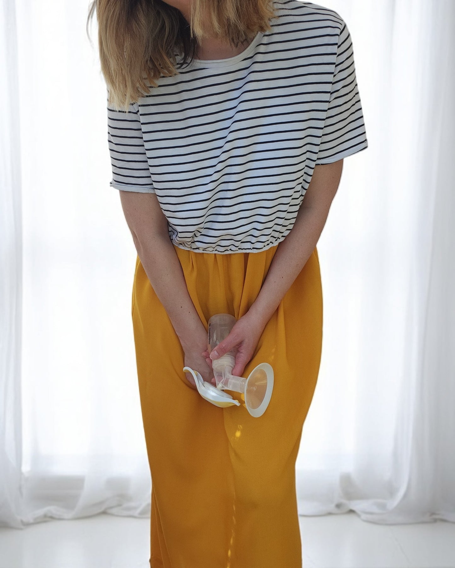 A blonde woman with shoulder-length hair, dressed in a touched out mama Upcycled Breastfeeding & Pumping Stripe Ochre Maxi Dress M/L, stands slightly bent forward holding a breast pump. Sheer white curtains in the background allow bright natural light to filter through.