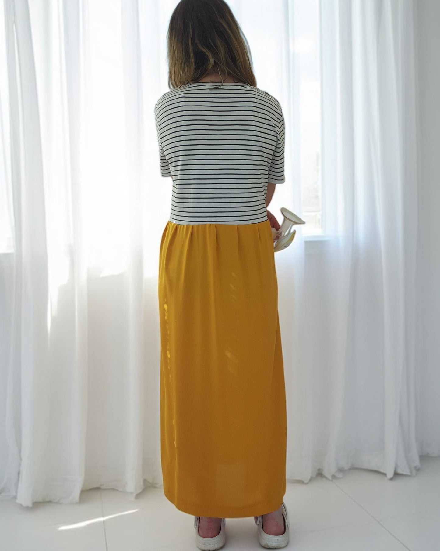 A person stands by white curtains, holding a ceramic cup. They wear the Upcycled Breastfeeding & Pumping Stripe Ochre Maxi Dress from touched out mama. Sunlight filters through, creating a soft and bright atmosphere.