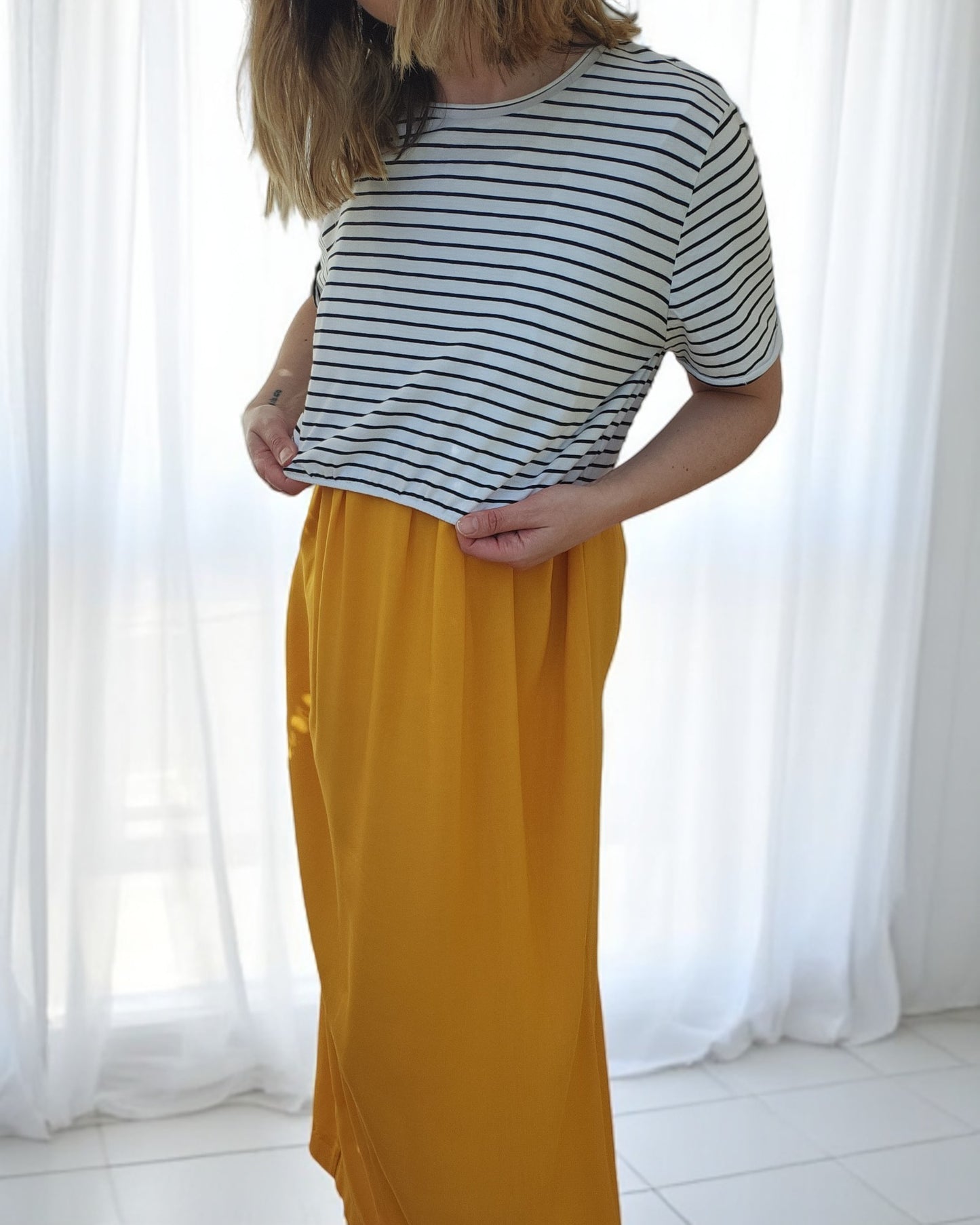 In a softly lit room, an individual models the Upcycled Breastfeeding & Pumping Stripe Ochre Maxi Dress from touched out mama, exemplifying eco-friendly fashion with a striped design paired beautifully with the ambient white curtains.