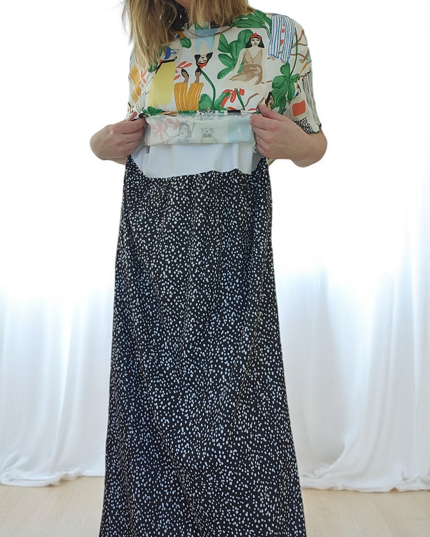 A person features an upcycled breastfeeding & pumping ZARA Tropical Maxi Dress by touched out mama layered over a black and white polka dot dress. Soft, white drapery in the background adds elegance while the unseen face highlights the unique texture and style blend.