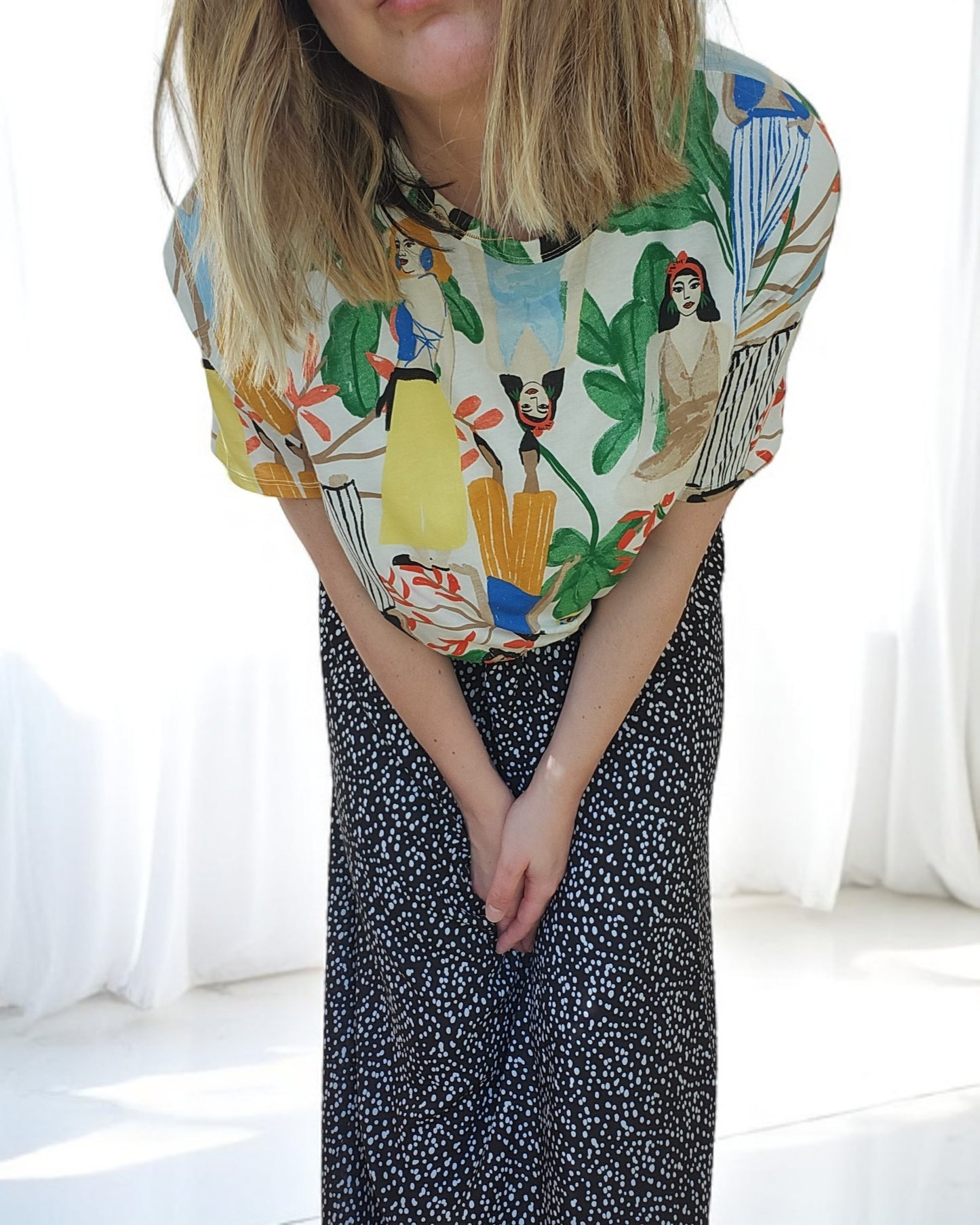 The woman with shoulder-length blonde hair dons an artistic Zara Tropical Maxi Dress by touched out mama. Leaning slightly forward with her hands clasped, she stands against a backdrop of bright curtains.