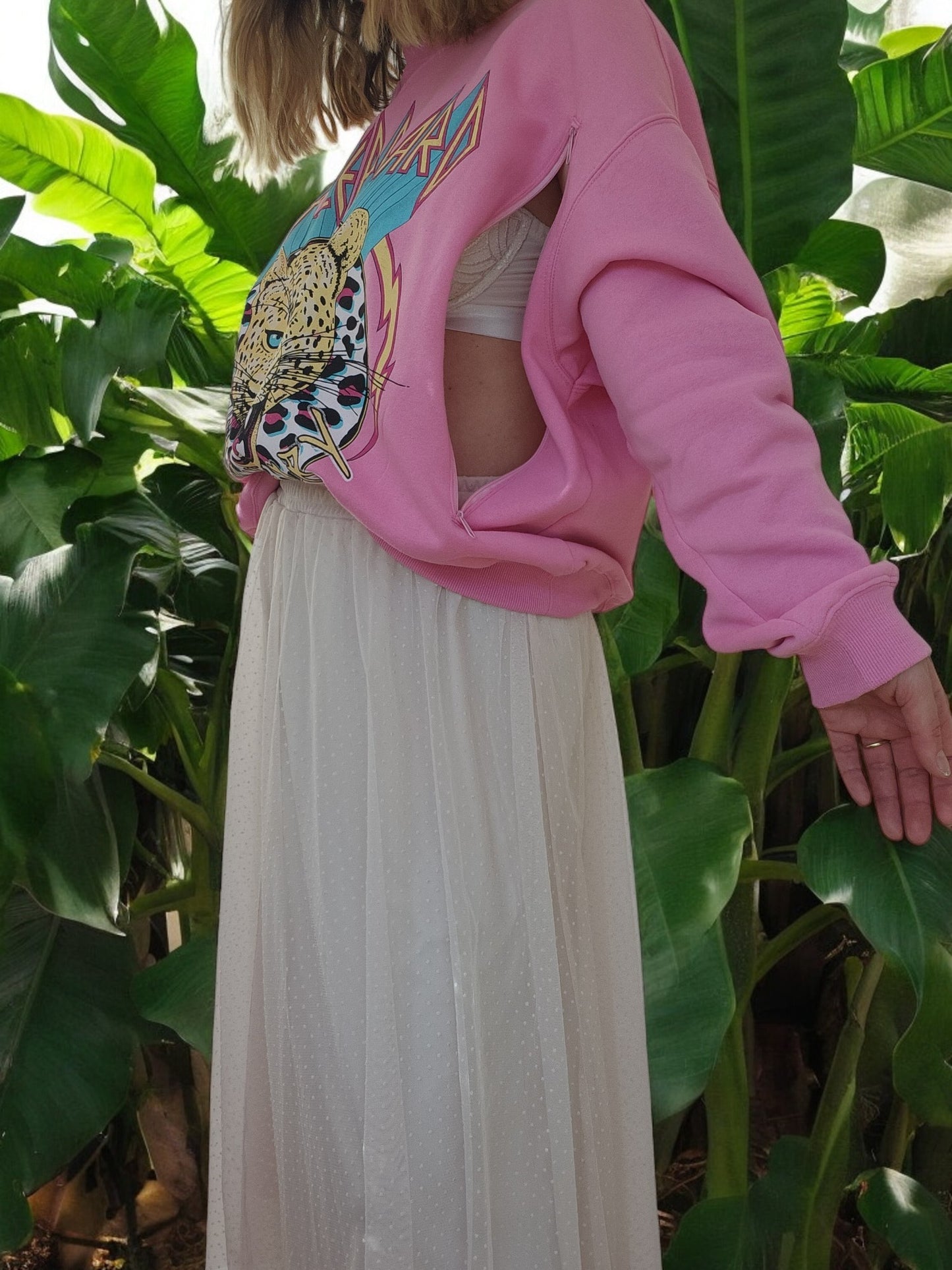 A woman stands among lush green leaves wearing an H&M Upcycled Breastfeeding Pink Def Leppard Band Sweatshirt (M) over a white pleated skirt and top—a chic look featuring sustainable nursing clothing.