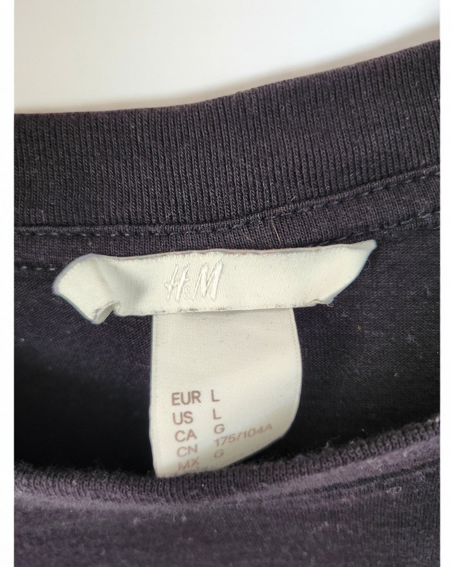 Close-up of a preloved H&M black t-shirt with a white label, size L for EU/US/CA, M for CN. The embroidered logo on the tag adds charm to this sustainably chic piece.