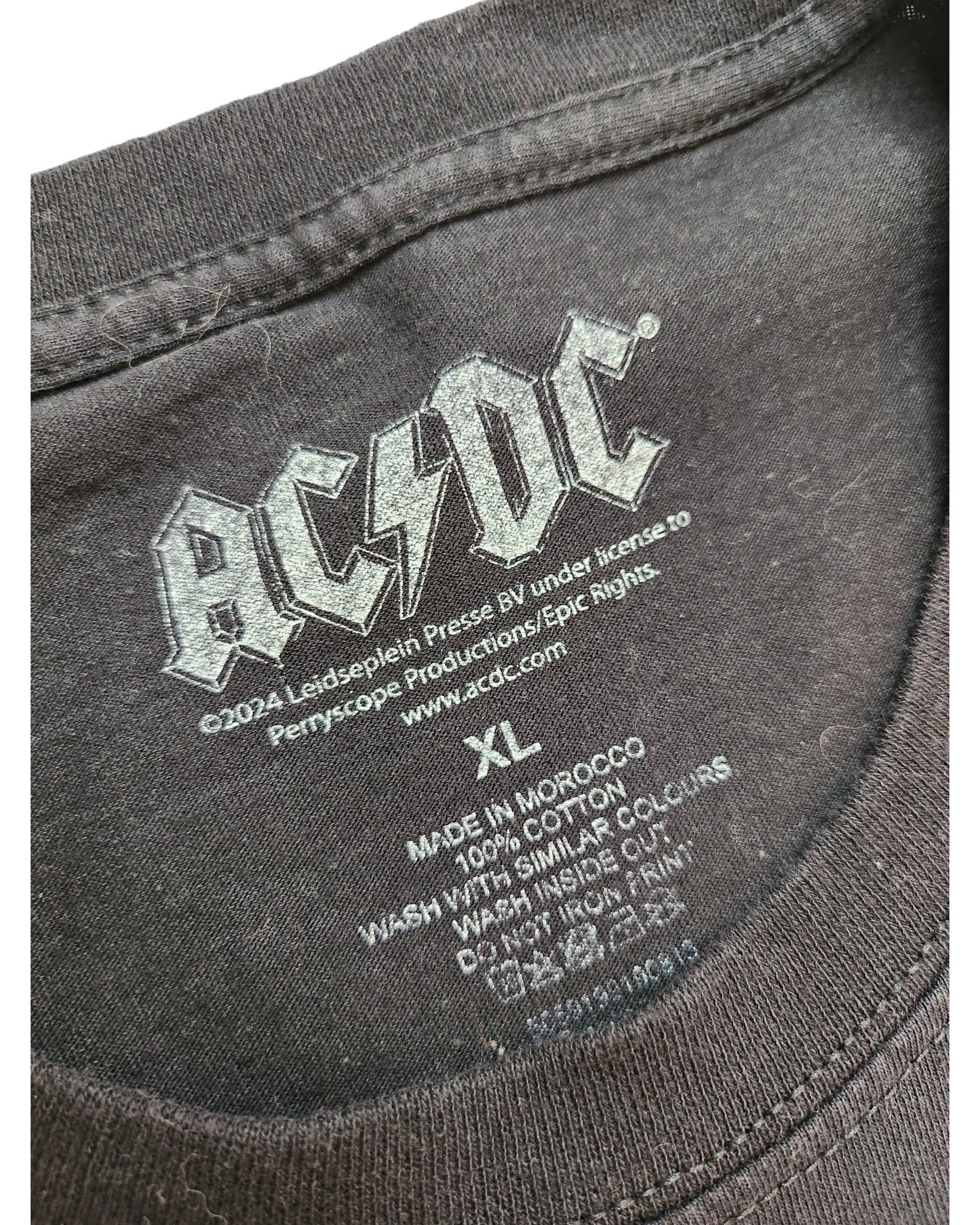 Close-up of a preloved touched out mama XL T-shirt label featuring an AC/DC logo, care instructions, and noting its made in Morocco from 100% cotton.