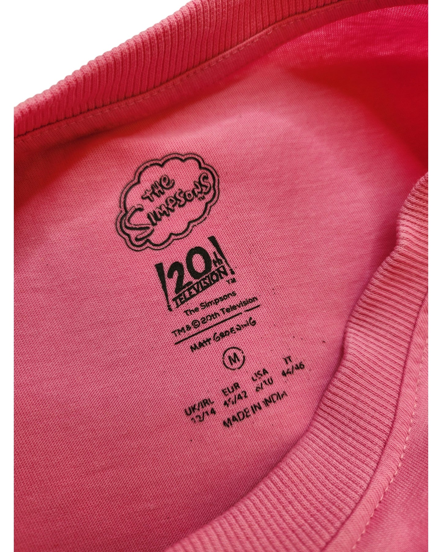 Close-up of a preloved Upcycled Breastfeeding & Pumping Lisa Simpson T-shirt (M) by touched out mama, highlighting its pink collar with black printed label info like size, various sizing systems, and Made in India; a piece rich with cherished history.