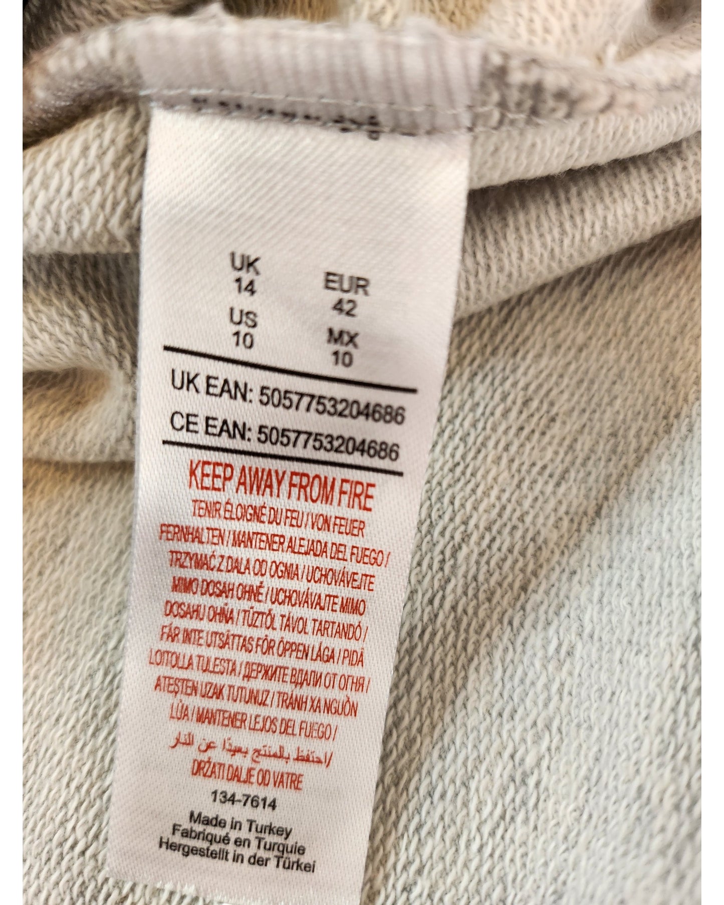 Close-up of a label on the touched out mama Upcycled Breastfeeding & Pumping Minnie & Mickey Sweatshirt UK14, showing size: UK (14), US (10), EUR (42), MX (10). It warns Keep Away From Fire in multiple languages and states its made in Turkey.