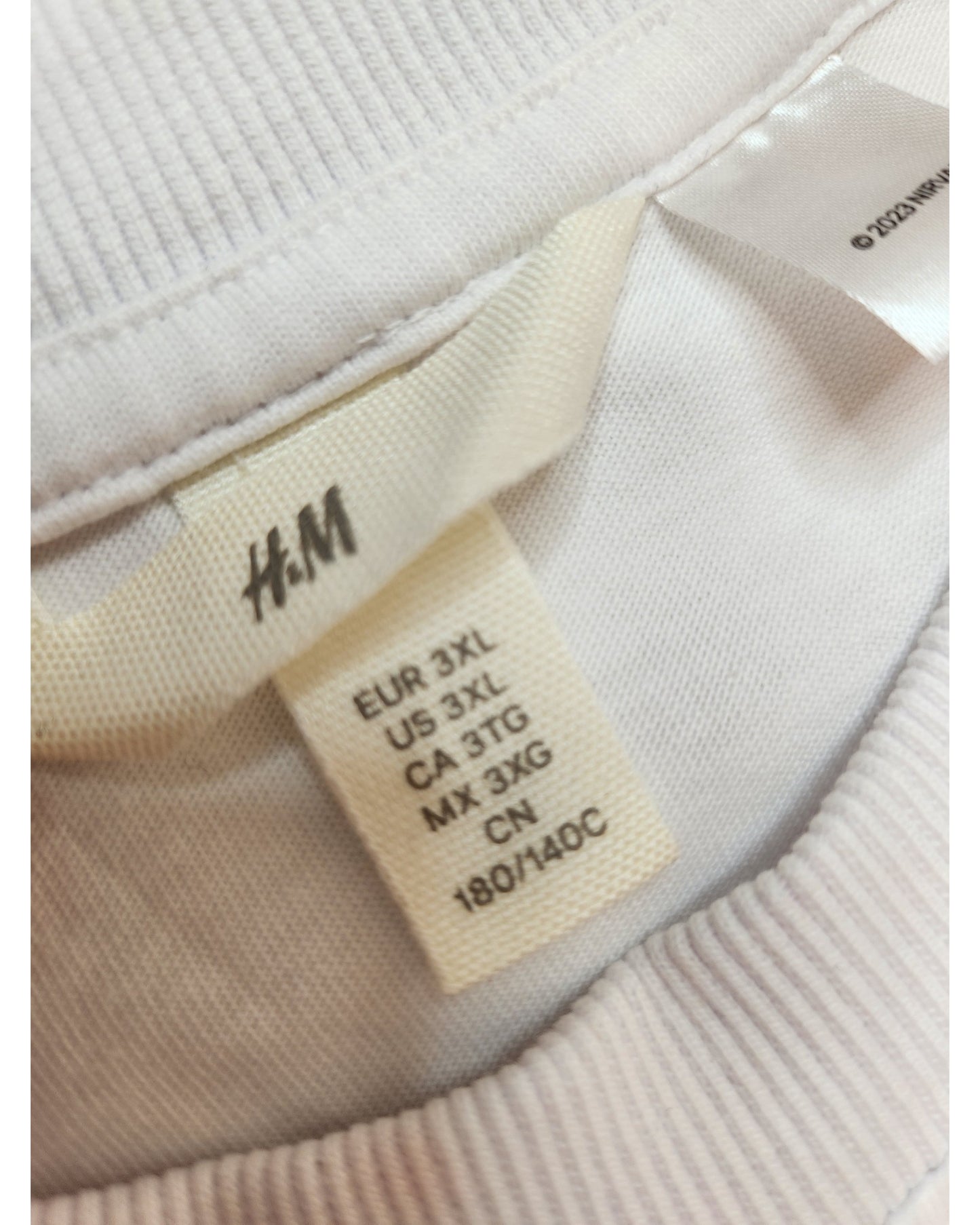 Close-up of a white clothing label from touched out mamas Upcycled Breastfeeding & Pumping White Nirvana T-shirt in size 3XL, showing sizes: EUR 3XL, US 3XL, CA 3TG, MX 3XG, and CN 180/140C. The label is sewn onto the white garment.