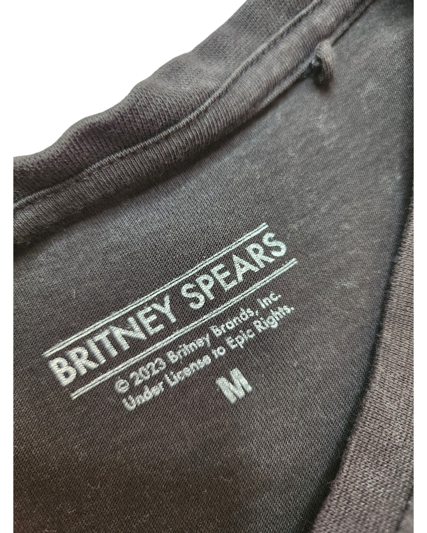 Close-up of a garment label on preloved clothing shows touched out mama, featuring Britney Spears and © 2022 Britney Brands, Inc. Brand details and licensing info are present along with size M, hinting at sustainable fashion.