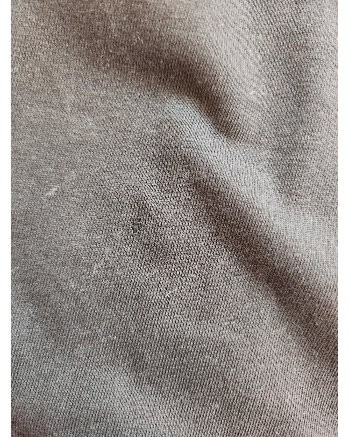 Close-up of the gray fabric from the Upcycled Breastfeeding & Pumping Britney T-shirt M by touched out mama, featuring a small hole. The textured, slightly creased, and faded areas lend character typical of preloved clothing to this unique piece.