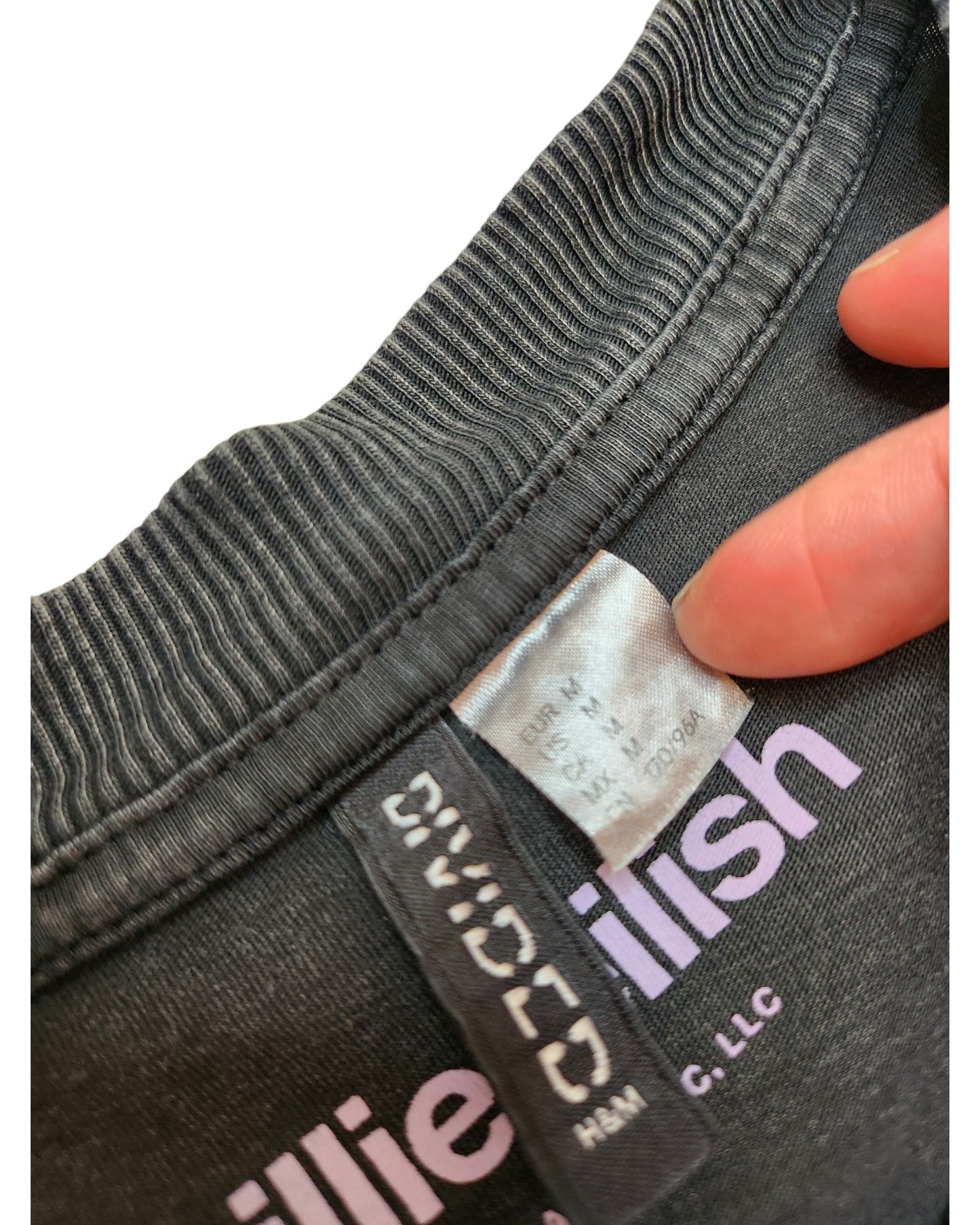 A close-up shows a black upcycled breastfeeding & pumping Billie Eilish T-shirt (EUR M, US M, CA M, CN 165/92A) by touched out mama. A label is held revealing the size tag as pink text design peeks through, highlighting its eco-friendly and sustainable style.