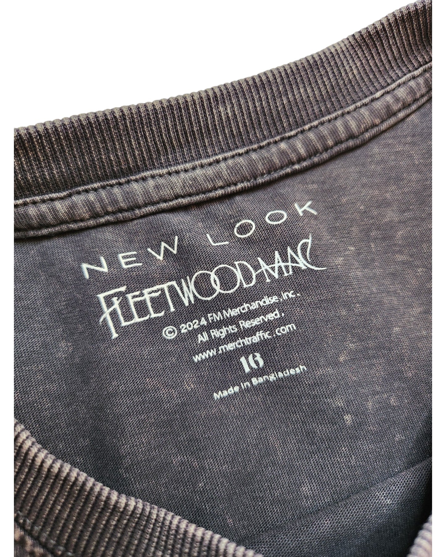 Close-up of a clothing label inside a dark garment. It features touched out mama above Fleetwood Mac, with 2024 copyright and merchtfx.com, indicating the product is an upcycled breastfeeding/pumping t-shirt made in Bangladesh from eco-friendly materials, size UK16.