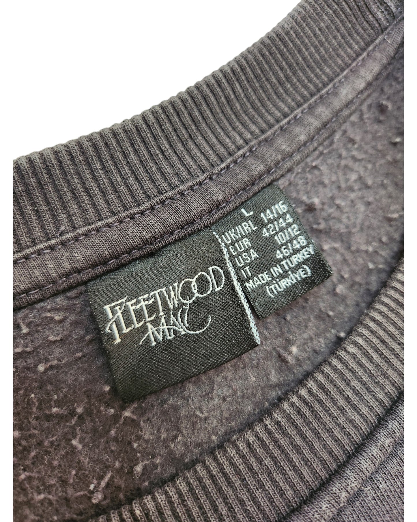 Close-up of a black label inside a dark grey Primark Upcycled Breastfeeding & Pumping Fleetwood Mac Floral Sweatshirt (L/UK14/16), showing the band’s logo, size info for various regions, and Made in Turkey at the bottom.