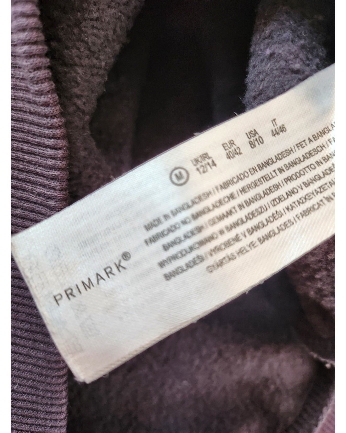 A close-up of a white Primark label on an upcycled dark grey Pink Floyd nursing & pumping sweatshirt (M, UK12/14), showing size info for different regions and care instructions—ideal for preloved fashion fans.