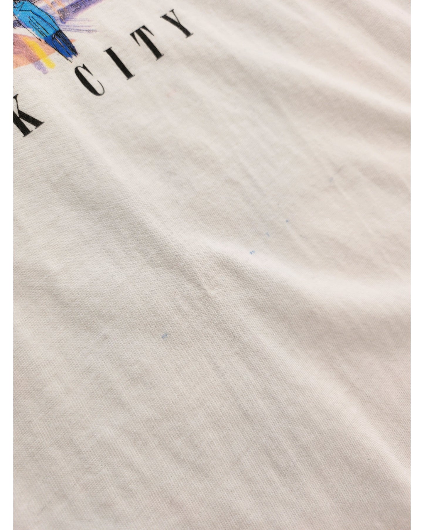 Close-up of a white Sun Wear Upcycled Breastfeeding & Pumping Times Square t-shirt (L) made from upcycled clothing, featuring faint blue ink marks, partial black CITY text, and a hint of colorful graphic in the upper left corner.