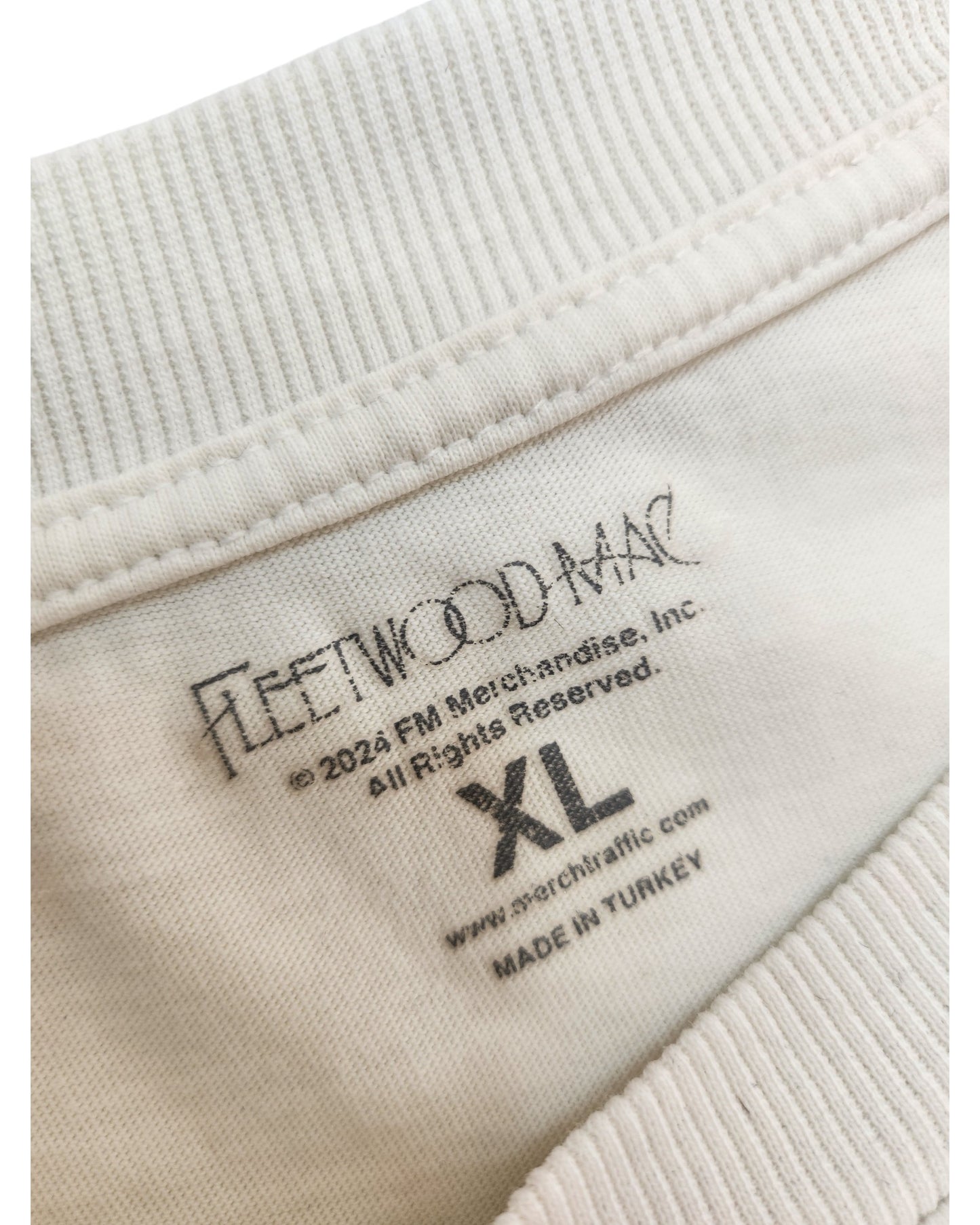 Close-up of a white clothing tag inside an upcycled breastfeeding & pumping Fleetwood Mac t-shirt (XL, white/black), made in Turkey by FM Merch-Mandise, Inc. Label lists brand as Fleetwood Mac.