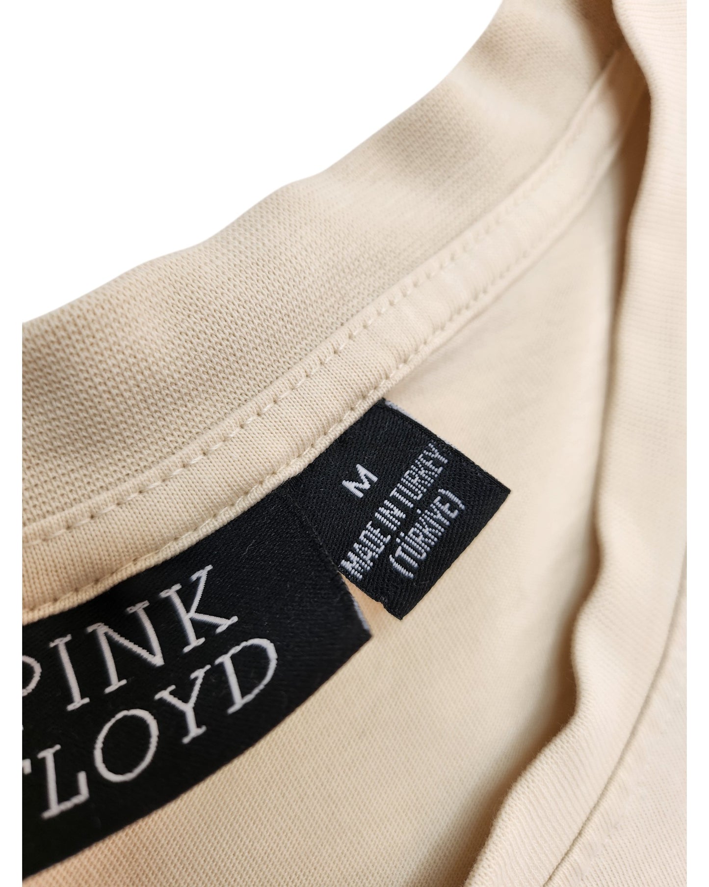 Close-up of a beige upcycled Pink Floyd breastfeeding & pumping T-shirt (size M) by Primark, featuring two black tags—one with the band’s name and one with “Made in Turkey” in English and Turkish. Ideal for fans of unique, sustainable fashion.