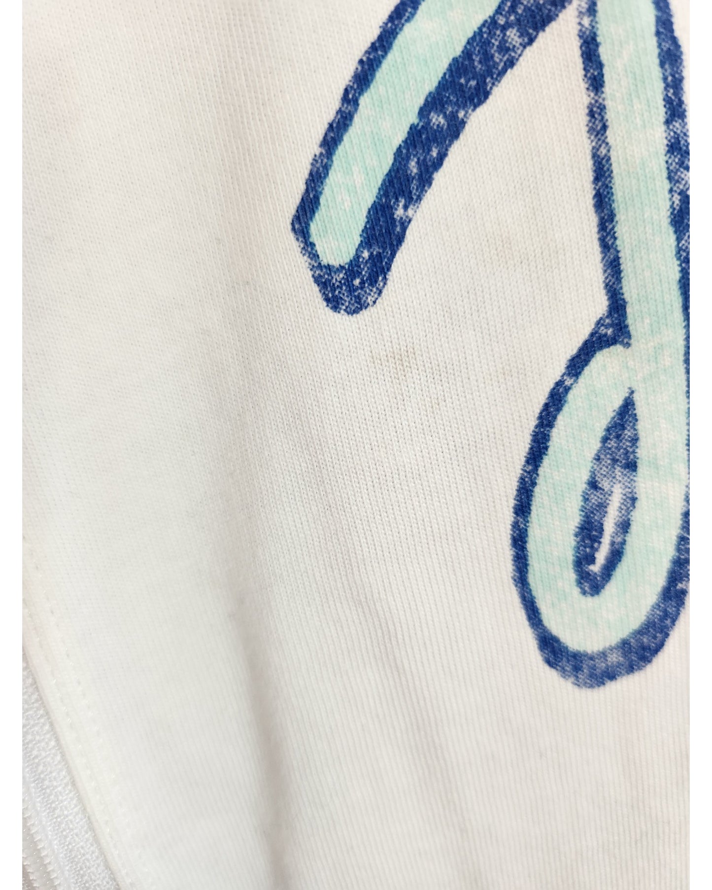 A close-up of an upcycled H&M Rolling Stones white/blue nursing & pumping t-shirt (L) with a large, blue-outlined symbol. The print is slightly worn and subtle stains are visible on the fabric.