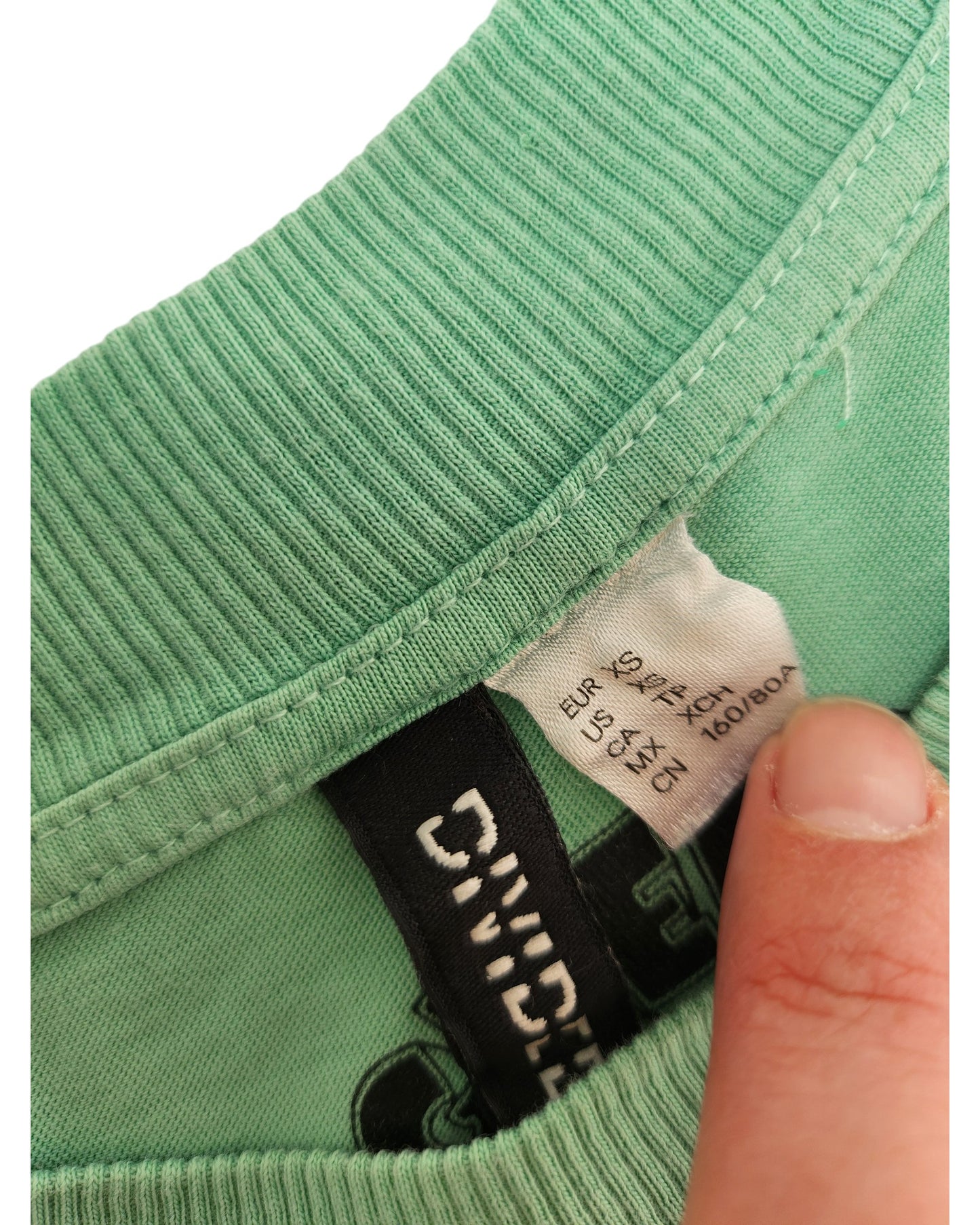 Close-up of a hand holding the collar of an H&M Upcycled Breastfeeding & Pumping Green Day Band t-shirt (XS), showing two labels with size info—including EUR S—and washing instructions in several languages.