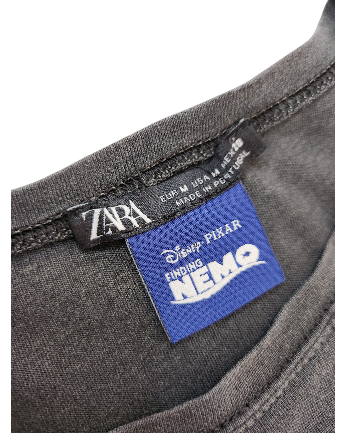 Close-up of the Upcycled Breastfeeding & Pumping ZARA Nemo t-shirt (size M) showing black ZARA and blue Disney Pixar Finding Nemo labels. Made in Portugal, this dark gray piece is part of our preloved fashion collection.