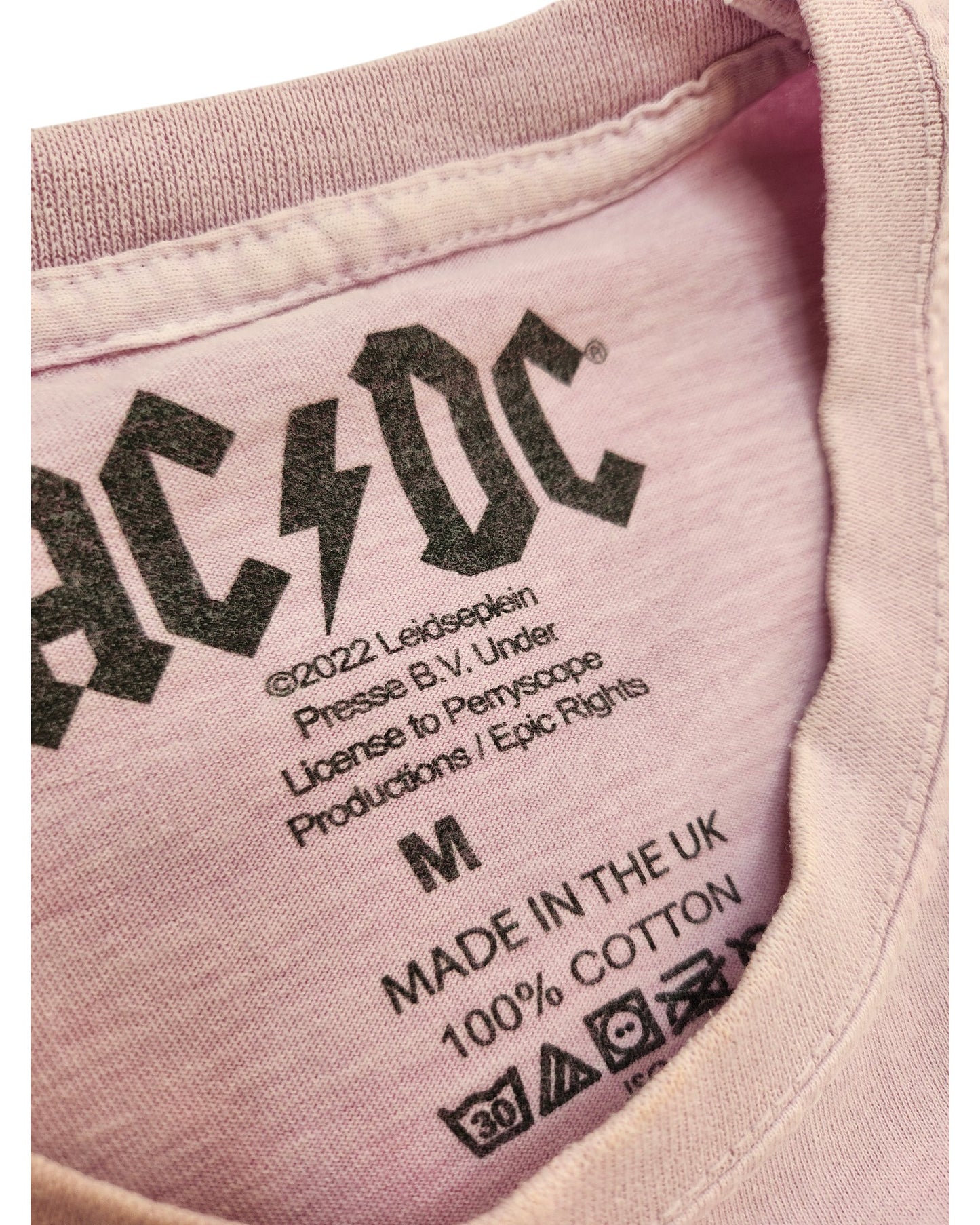 Close-up of a lilac AC/DC Upcycled Breastfeeding & Pumping T-shirt (size M) neck label showing logo, copyright, size, Made in UK, 100% cotton—crafted as eco-friendly apparel for conscious fans.