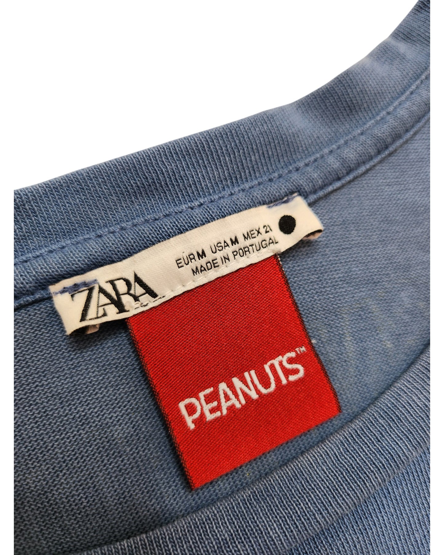 Close-up of a blue ZARA Snoopy t-shirt collar, showing a white ZARA label with size and Made in Portugal plus a red Peanuts™ tag—highlighting this unique upcycled breastfeeding & pumping top in size M.
