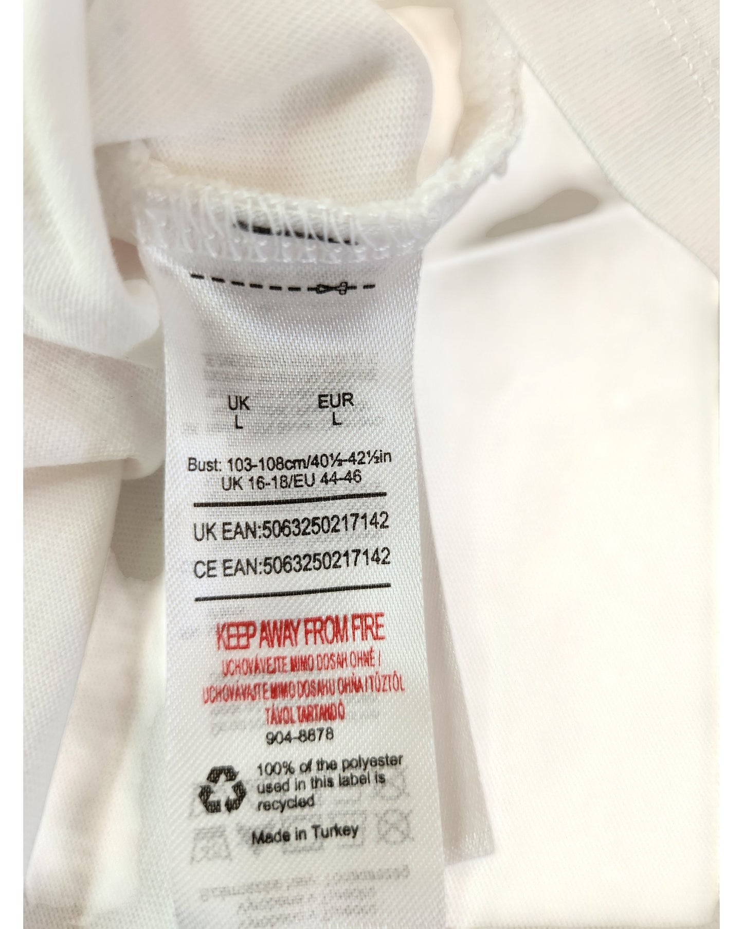 A close-up of a white label on the Upcycled Breastfeeding & Pumping Def Leppard t-shirt (L), showing size, care details, product codes, a red keep away from fire warning, and noting its made in Turkey from 100% polyester.