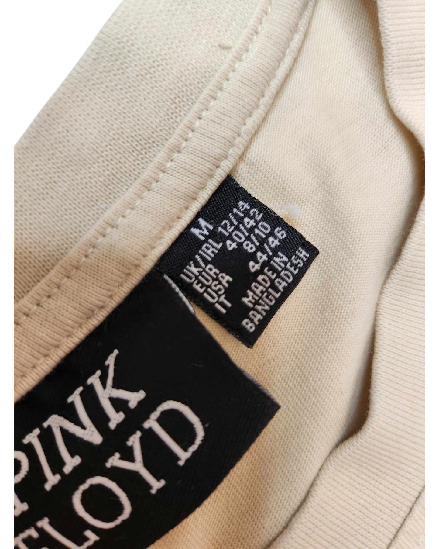 Close-up of a beige Primark t-shirt with two tags: one reads PINK FLOYD in white, the other shows size, material, and care info. This upcycled breastfeeding & pumping top (M/UK12-14) highlights eco-friendly fashion made in Bangladesh.