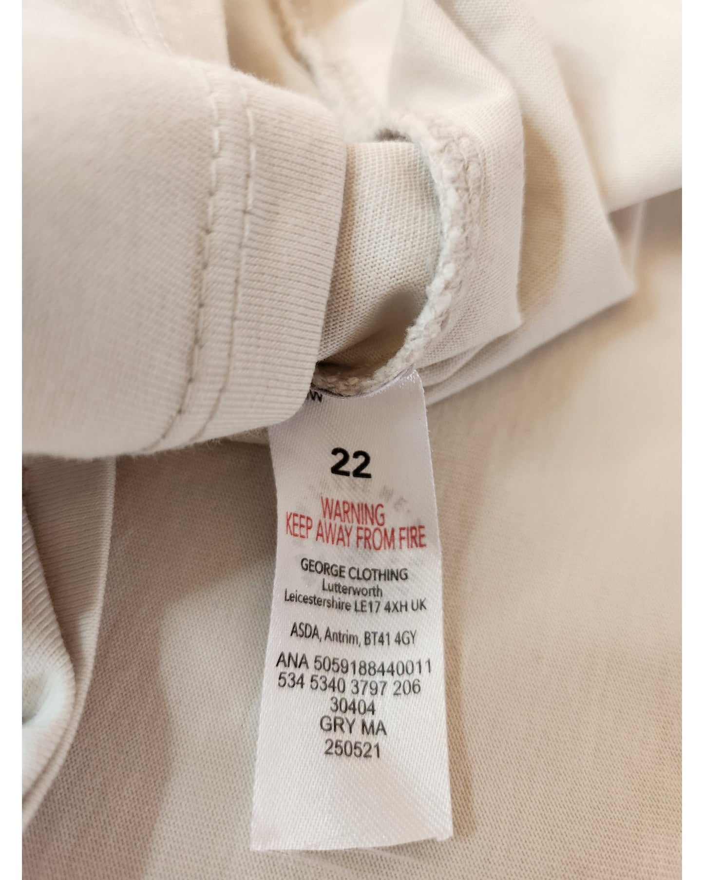 A white label on a light fabric for the Upcycled Breastfeeding & Pumping Cream Care Bears t-shirt UK22 by George shows size 22, a red KEEP AWAY FROM FIRE warning, brand details, barcodes, and eco-friendly fashion info.