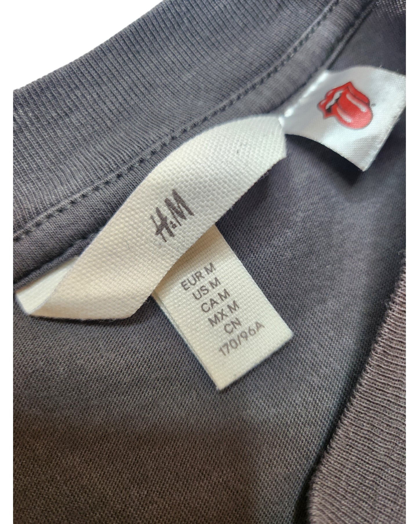 Close-up of a gray H&M Upcycled Breastfeeding & Pumping The Rolling Stones Yellow Stars Band t-shirt (size M), showing two tags: one with sizing info and the other featuring a small red lips graphic.