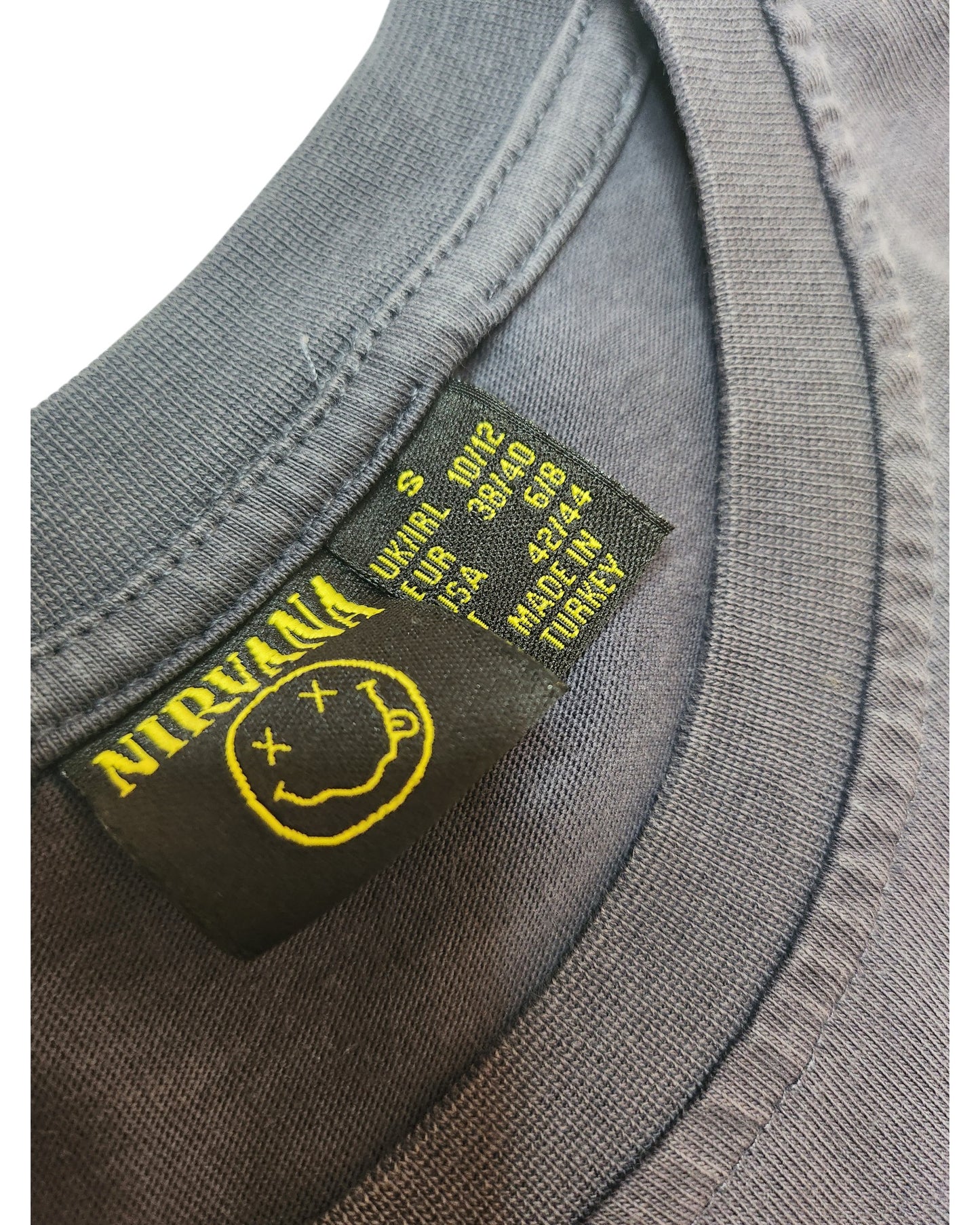 Close-up of a Primark Upcycled Breastfeeding & Pumping Yellow Smile Nirvana Band t-shirt (S, UK10/12) collar, showing black and yellow tag with the band’s name, smiley face logo, size small, and Made in Turkey detail.