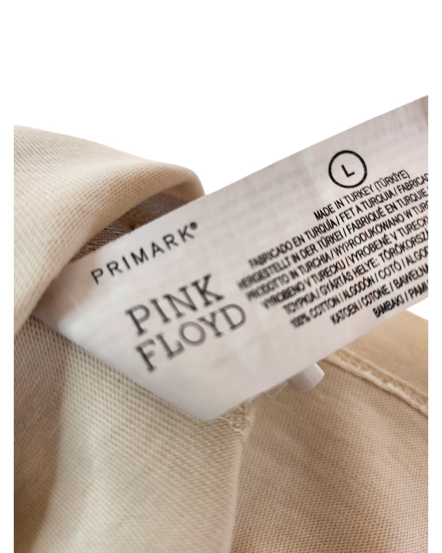 A close-up of a tag inside a beige, upcycled Primark breastfeeding & pumping t-shirt (Pink Floyd 1977 Band, size L), showing PRIMARK at the top, PINK FLOYD in bold, care info in various languages, and size L.